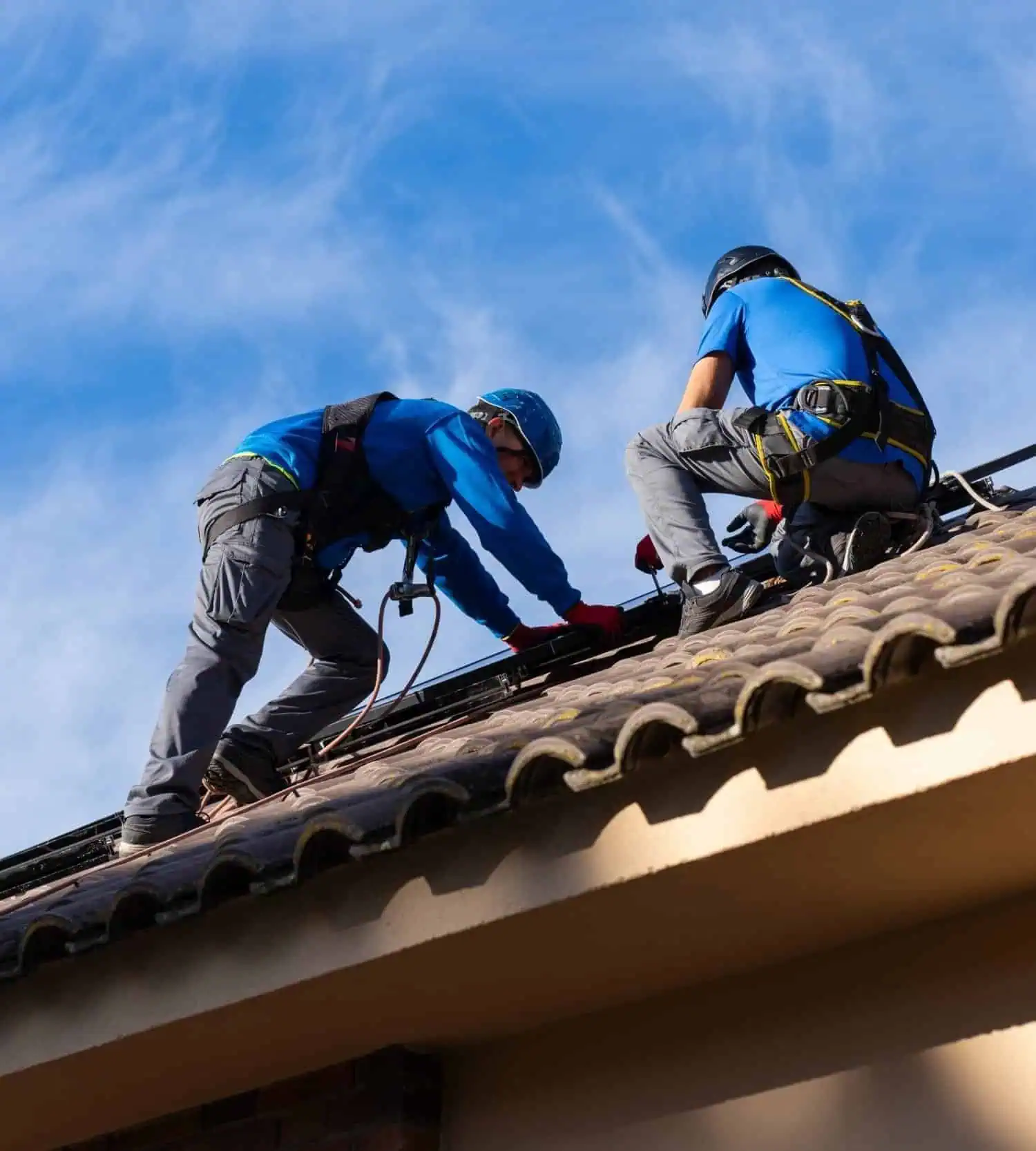 Two roofers working on a tiled roof with safety gear and harnesses.