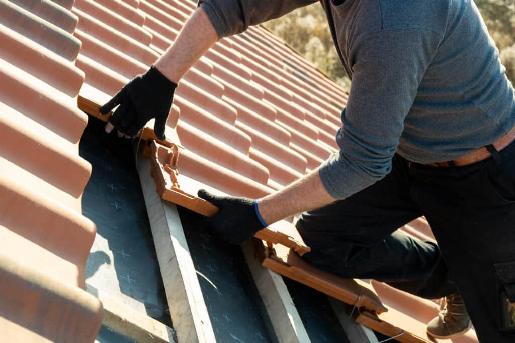 Roofing installer working on a tiled roof with tools and safety gloves.